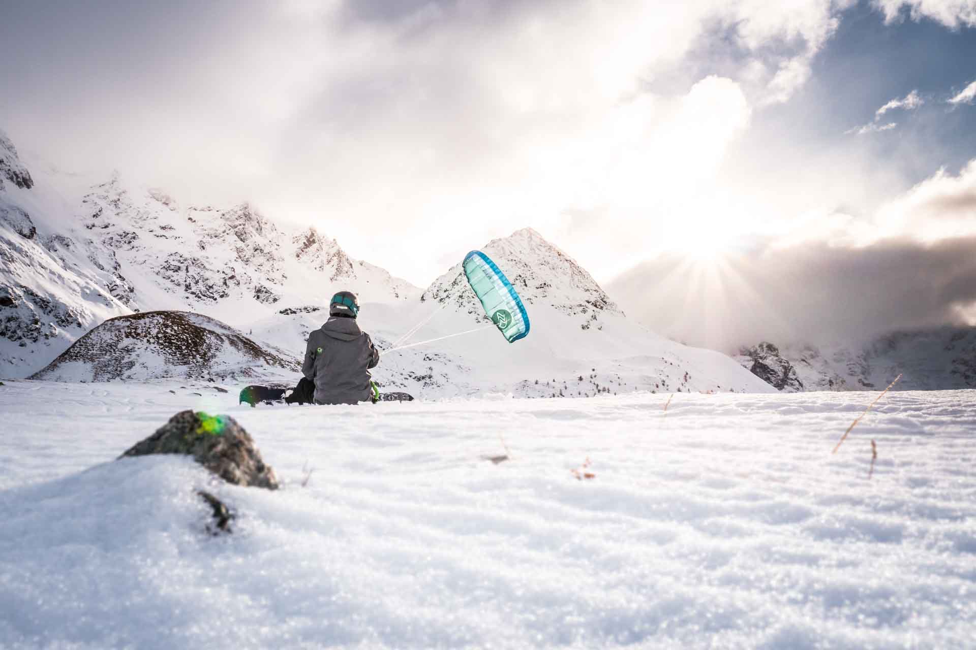 Snowboarder sitzt mit Kite am Berg und blickt richtung Sonne