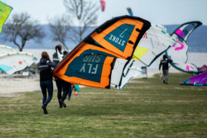 Die Teilnehmer der LakeUnited Academy gehen mit aufgebauten Schirmen gemeinsam Richtung Wasser zum Kitesurfen.
